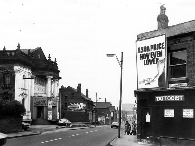 A tattooist on the corner of Domestic Street offers 'expert tattoo removal' and 'painless ear piercing'. An advertising hoarding for Asda supermarket covers the wall. On the left of the picture is Holbeck Mills Cash and Carry Carpet Warehouse, formerly the United Methodist Chapel. The junctions with Ingram View and Willoughby Terrace are at either side.