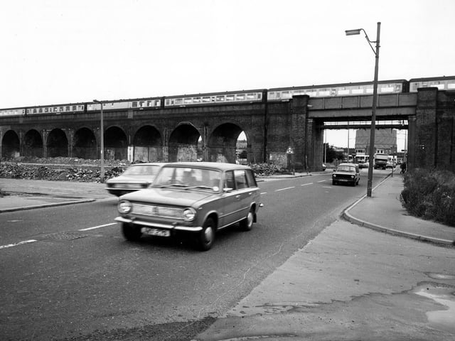 Tthe railway viaduct crossing Domestic Street in February 1980. An Inter-City train with many carriages is passing overhead. Cars can be seen on the road. A gasometer is visible in the distance through the archway.