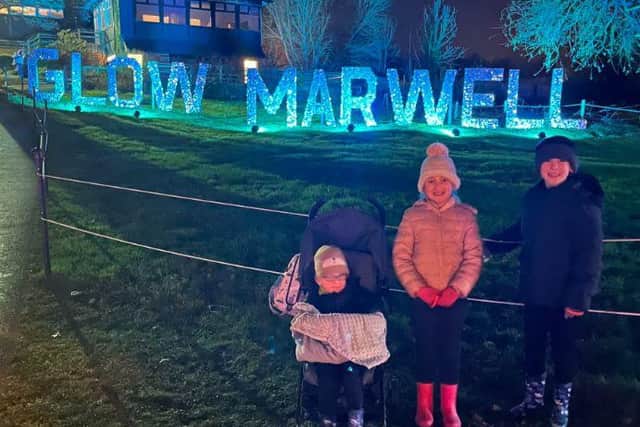 Three children enjoy an evening at Glow Marwell light trail, with one child in a stroller and two standing beside, smiling in warm coats and boots under glowing blue lights.