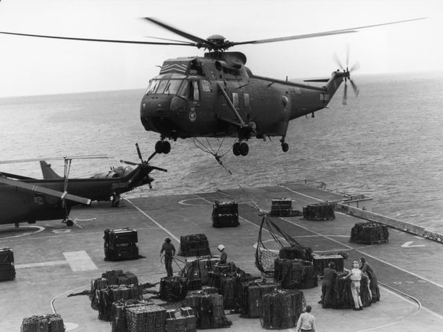 A Sea King helicopter hovering above HMS Hermes to airlift ammunition for redistribution to other ships in the convoy, April 1982.