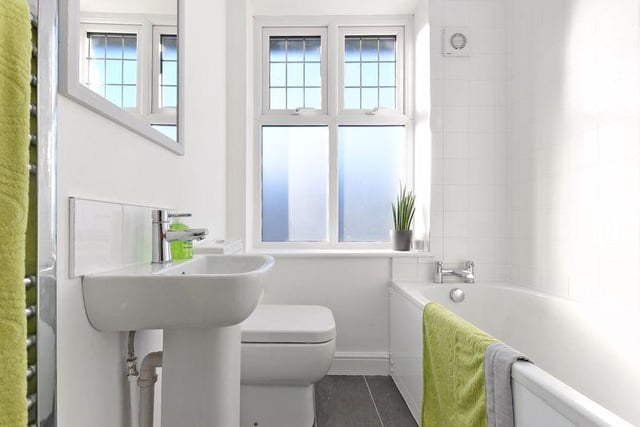 The bathroom has a white suite, a bath with shower over and glass shower screen, WC, sink and chrome towel radiator, white tiled walls and grey tiled floors.