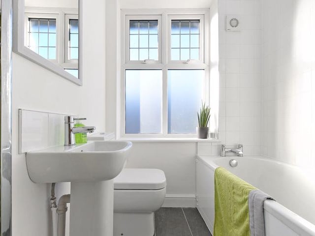 The bathroom has a white suite, a bath with shower over and glass shower screen, WC, sink and chrome towel radiator, white tiled walls and grey tiled floors.