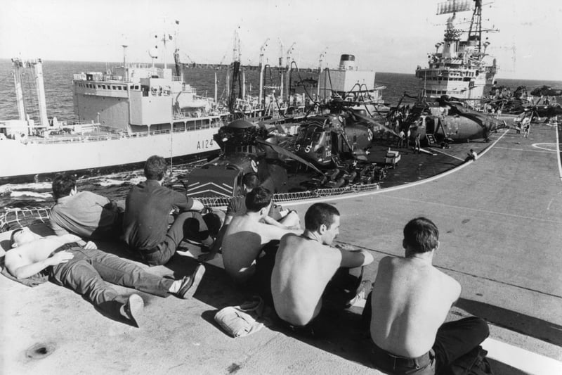 Marines relaxing on the 'ski jump' ramp of HMS Hermes, as she is replenished at sea by a Royal Fleet auxiliary vessel, on the way to the Falkland Islands in 1982. Westland Sea King helicopters can be seen on deck.