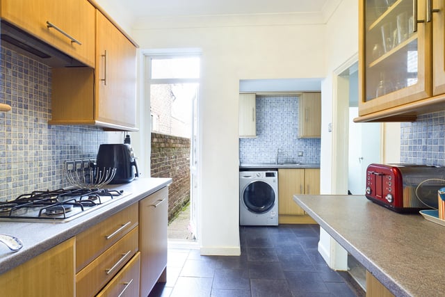 The galley style kitchen has modern beech style units, integrated oven, hob and extractor with space for a washing machine and blue mosaic tiling.