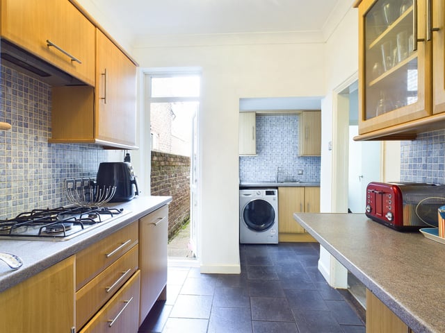 The galley style kitchen has modern beech style units, integrated oven, hob and extractor with space for a washing machine and blue mosaic tiling.