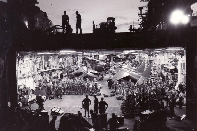 Dawn breaks over HMS Hermes en route to the South Atlantic from Portsmouth in April 1982. Royal Marines line up for a weapons check in the brightly-lit hangar of the task force flagship. In the background are tightly-packed Sea Harriers and Sea King helicopters, while in the foreground, shadowy figures of sailors wait with boxes of ammunition.