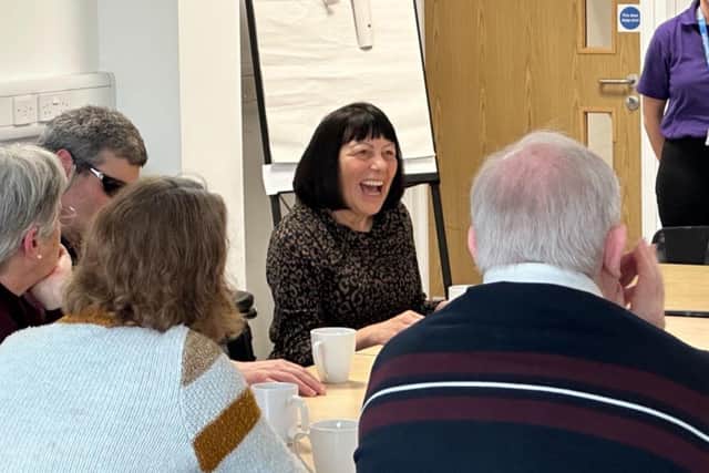 A group of people sit around a table with mugs, engaged in lively conversation. A woman in the centre is laughing warmly, creating a joyful atmosphere. A flipchart and door are visible in the background. Picture: OpenSight