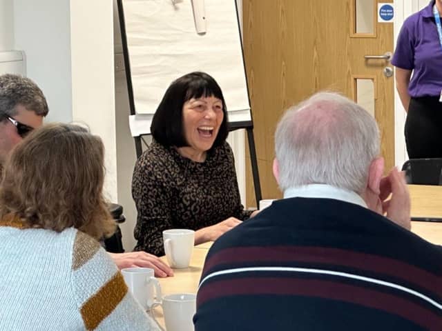 A group of people sit around a table with mugs, engaged in lively conversation. A woman in the centre is laughing warmly, creating a joyful atmosphere. A flipchart and door are visible in the background. Picture: OpenSight