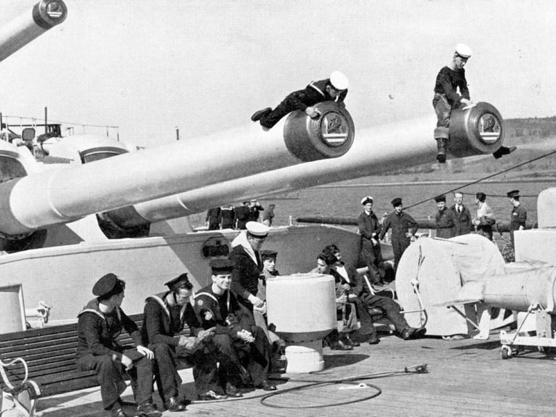 The ship's company of the battleship HMS Vanguard. The two men on the barrels of A turret are polishing the muzzle and the tampions that can be seen inserted into the barrels.