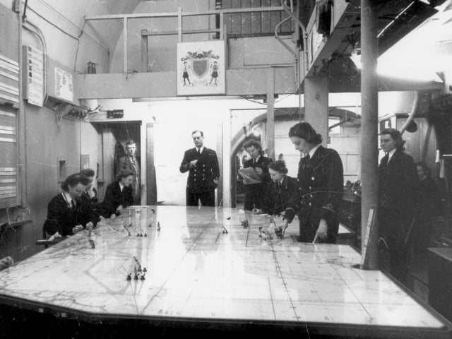 The plotting room in the underground headquarters at Fort Southwick, 1944. Tunnels beneath the fort housed the Combined Operations Underground HQ which played an important command and communications role in the D-Day Landings. In the plotting room, Wrens (members of the Women's Royal Naval Service) plotted naval movements in the English Channel on a map. In the background can be seen the unofficial coat of arms of the plotting room. The coat of arms was designed by Wren 3rd Officer Dorothy C.P. Ward who worked on the main plot, and is now on display at The D-Day Story. It is believed that the artist was Dorothy Cordelia Phyllis Ward (1909-1994). She was born in  Plymouth and did an art degree at the Royal College of Art in 1933. In later life she lived in Tavistock, Devon, and was well-regarded for her paintings of local scenes. (Not to be confused with the actor of the same name who lived around the same time.)
