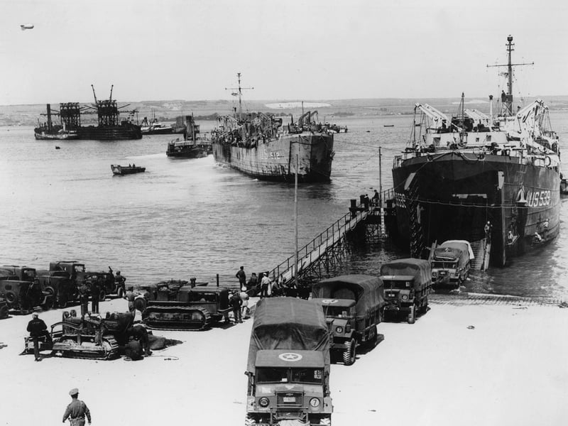 Vehicles bearing the Allied recognition star (they might be British or Canadian, not just US) reversing onto a Landing Ship, Tank (LST) at Hardway, Gosport. The LST is crewed by US Navy personnel. Another LST and a refuelling bare are in the left background.