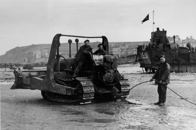 A bulldozer on a French beach (possibly Arromanches) with houses and landing craft in the background. "Store 6b, Depot Gold" is written on the rear of the bulldozer.