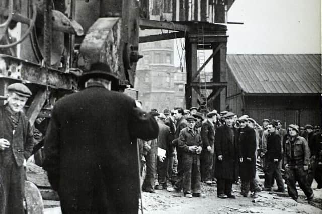A view of a construction site with a large concrete mixer in the right and a tower crane in the centre of the image.  In the centre foreground of the image is probably Winston Churchill.  A group of workers are gathering in front of him as he approaches.  The image probably shows an official visit to one of the Mulberry Harbour construction sites.