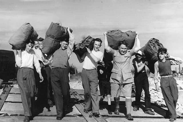 Tent-carrying  St Judes Boys' Club members, with George Harris in tropical shorts, walking down the boat ramp opposite the barracks to load the landing craft.