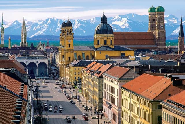 A panoramic view of Munich with the green domes of the Frauenkirche and the peaks of the Austrian Alps in the background.