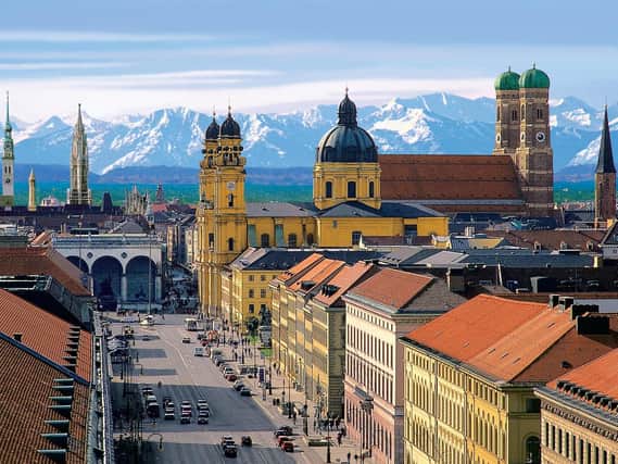 A panoramic view of Munich with the green domes of the Frauenkirche and the peaks of the Austrian Alps in the background.